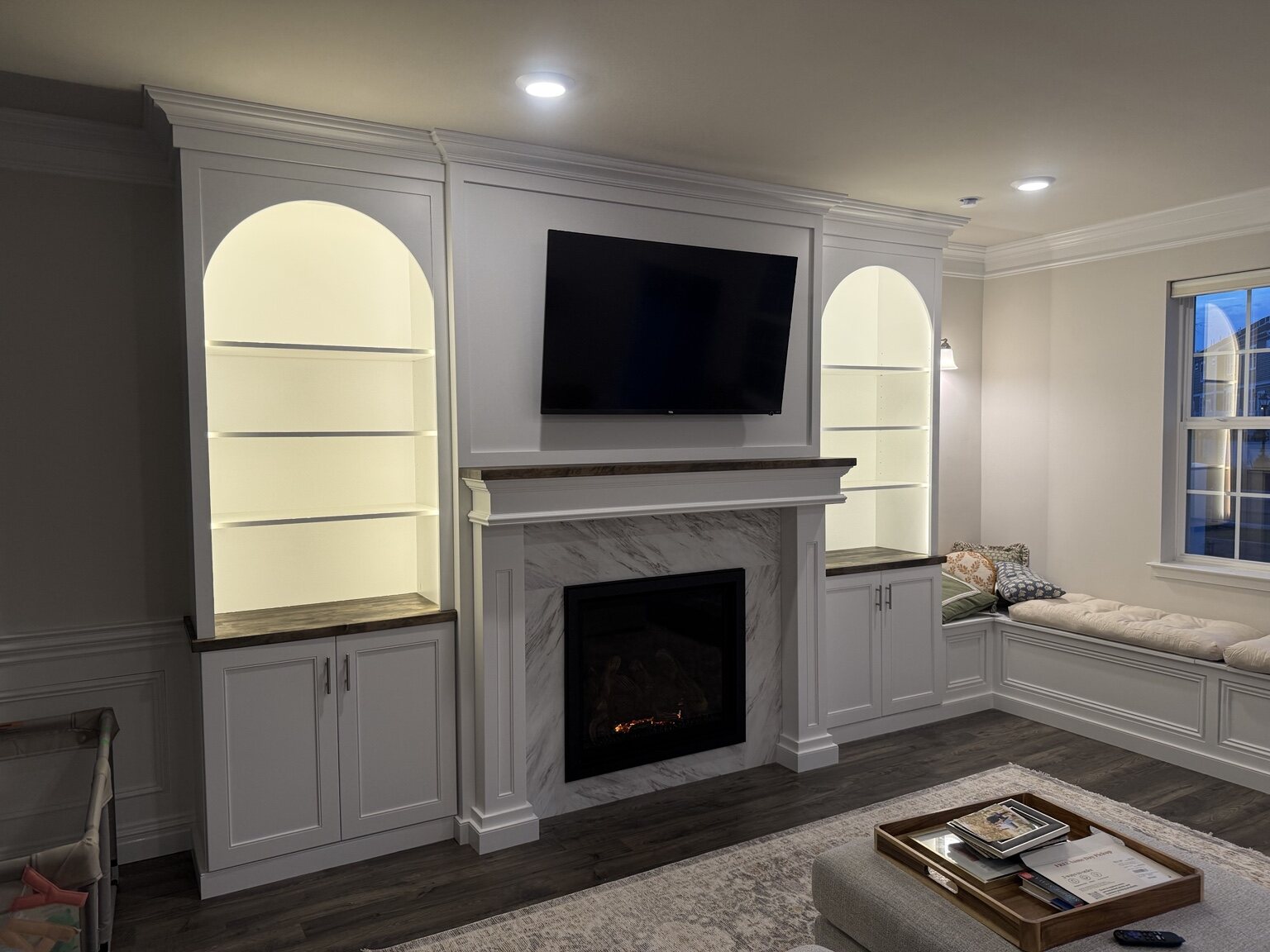 Custom fireplace wall with illuminated built-in shelving and window-seat cabinetry